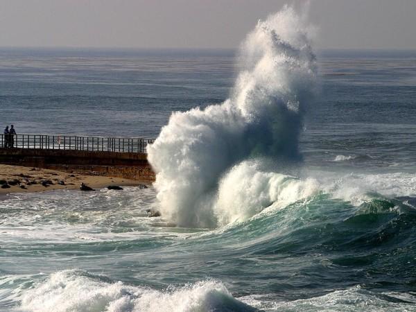 A huge wave crashes into rocks along the shoreline as beachgoers look on, marveling at the size of the waves and the height the water reaches as it splashes in every direction.