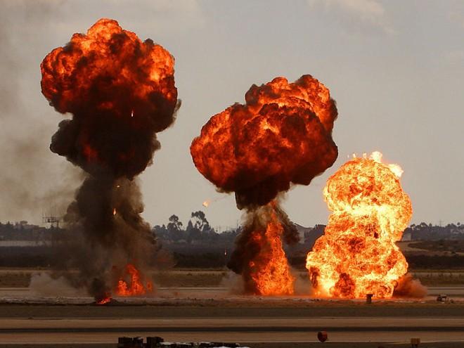 Three massive explosions take place side by side at an outdoor test facility. One after another the explosions burst smoke and giant flames high into the air.